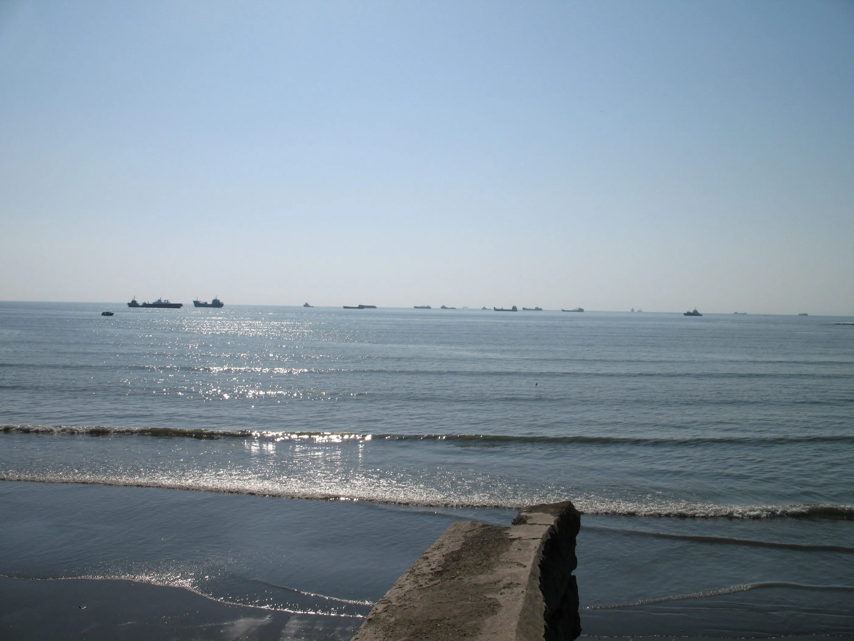 Ships at anchor on the horizon off a breakwater.