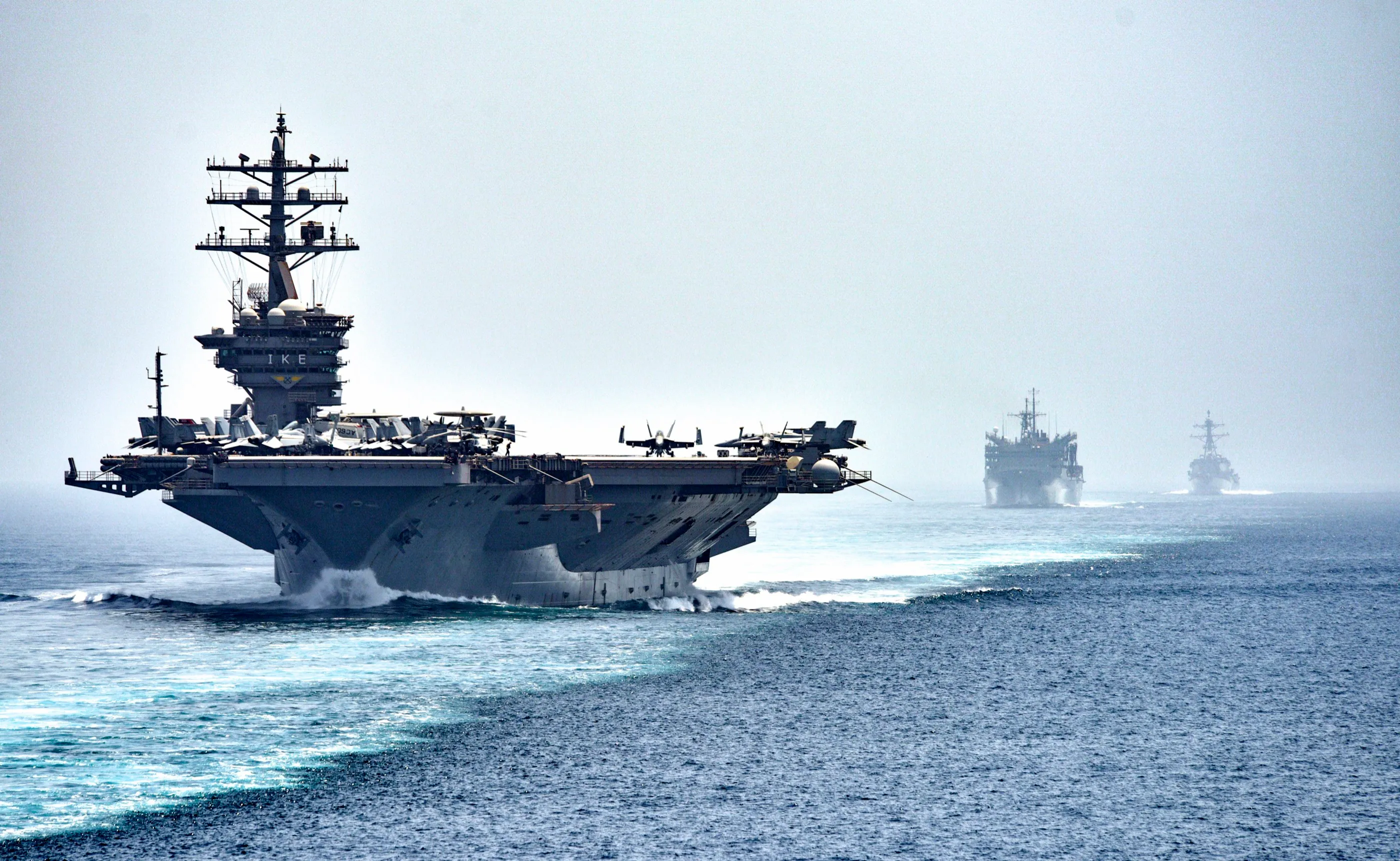 An aircraft carrier leads a supply ship and a destroyer through hazy water.