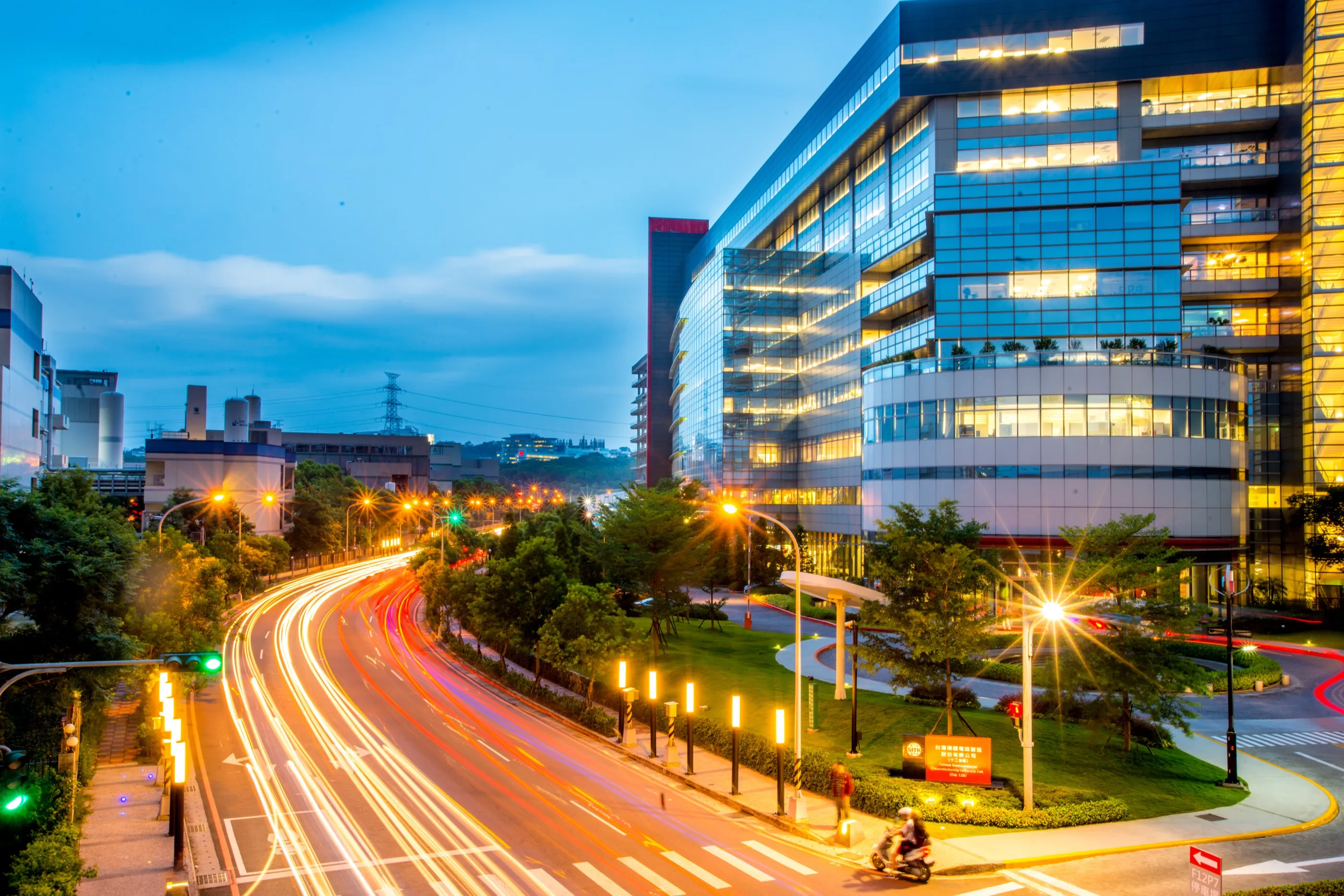 A glass-fronted factory building beside a road with light trails at dusk.