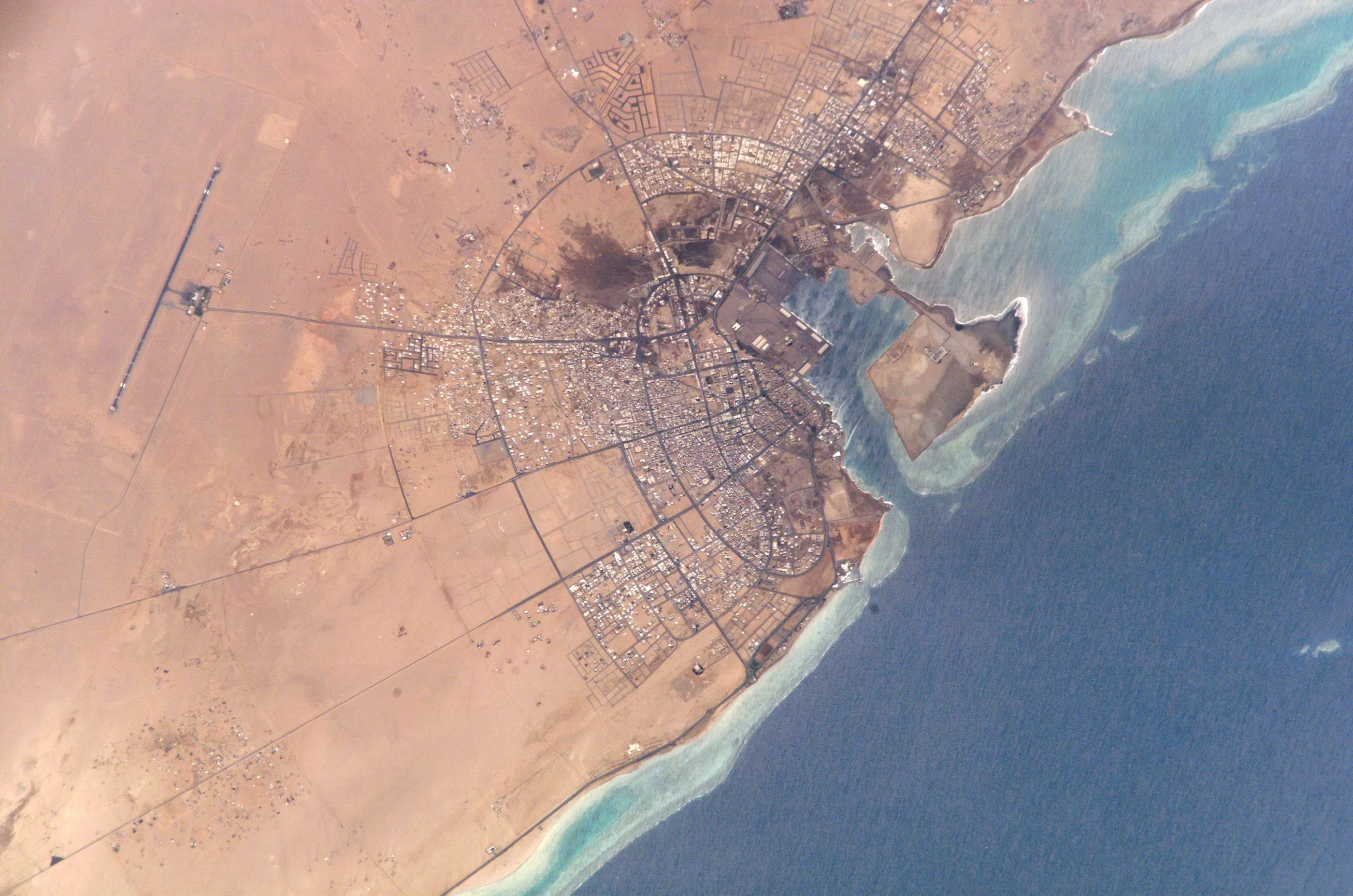 A desert coastline and port town beside the reefs of the Red Sea, seen from orbit.